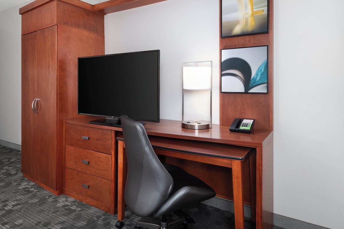 A hotel room with a wooden desk, a flat-screen TV, a desk lamp, a telephone, and a black office chair.