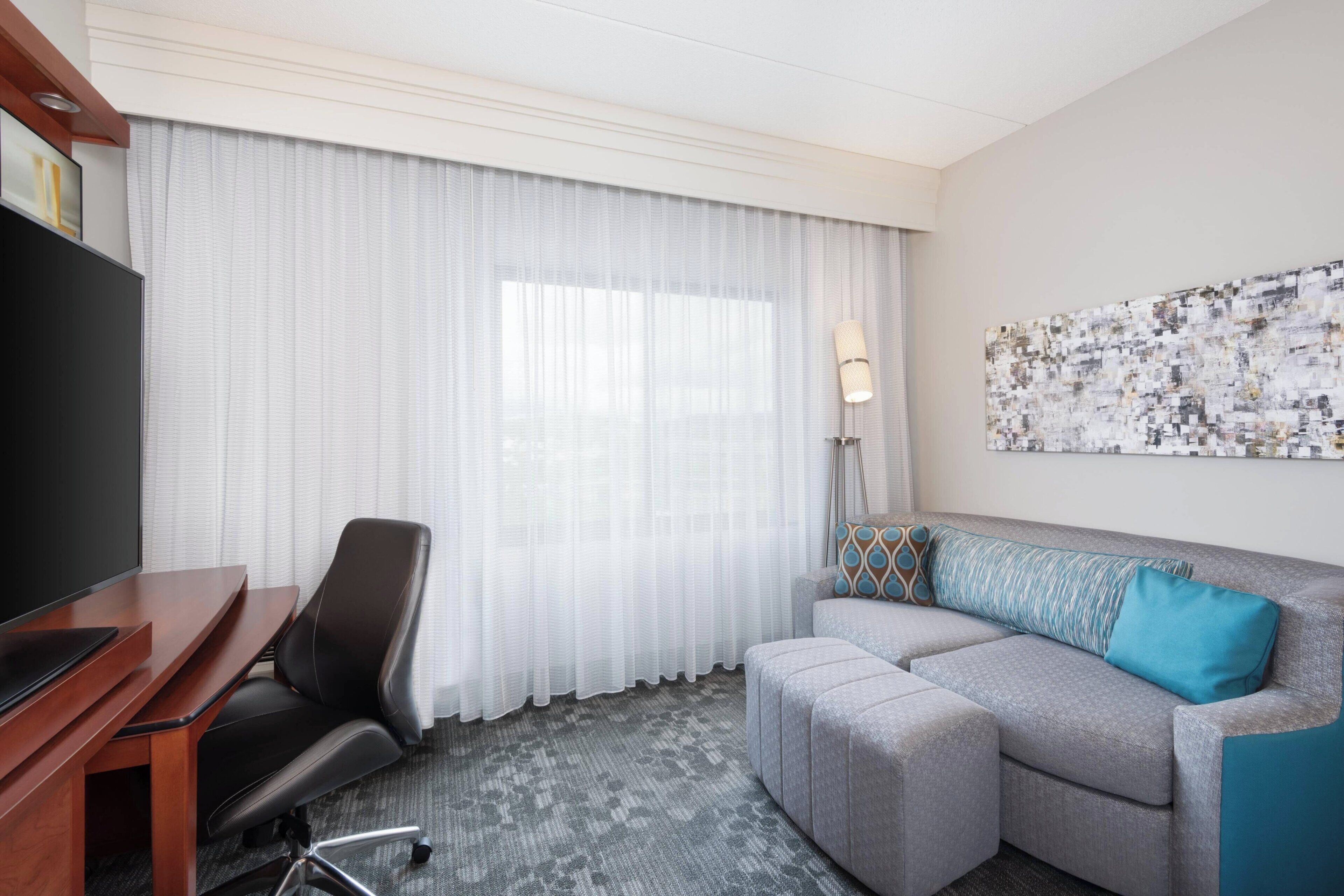 Une chambre d’hôtel équipée d’un téléviseur à écran plat, d’un bureau, d’un fauteuil de bureau noir, d’un canapé gris agrémenté d’un coussin bleu, d’un tapis à motifs et d’une grande fenêtre avec des rideaux légers.