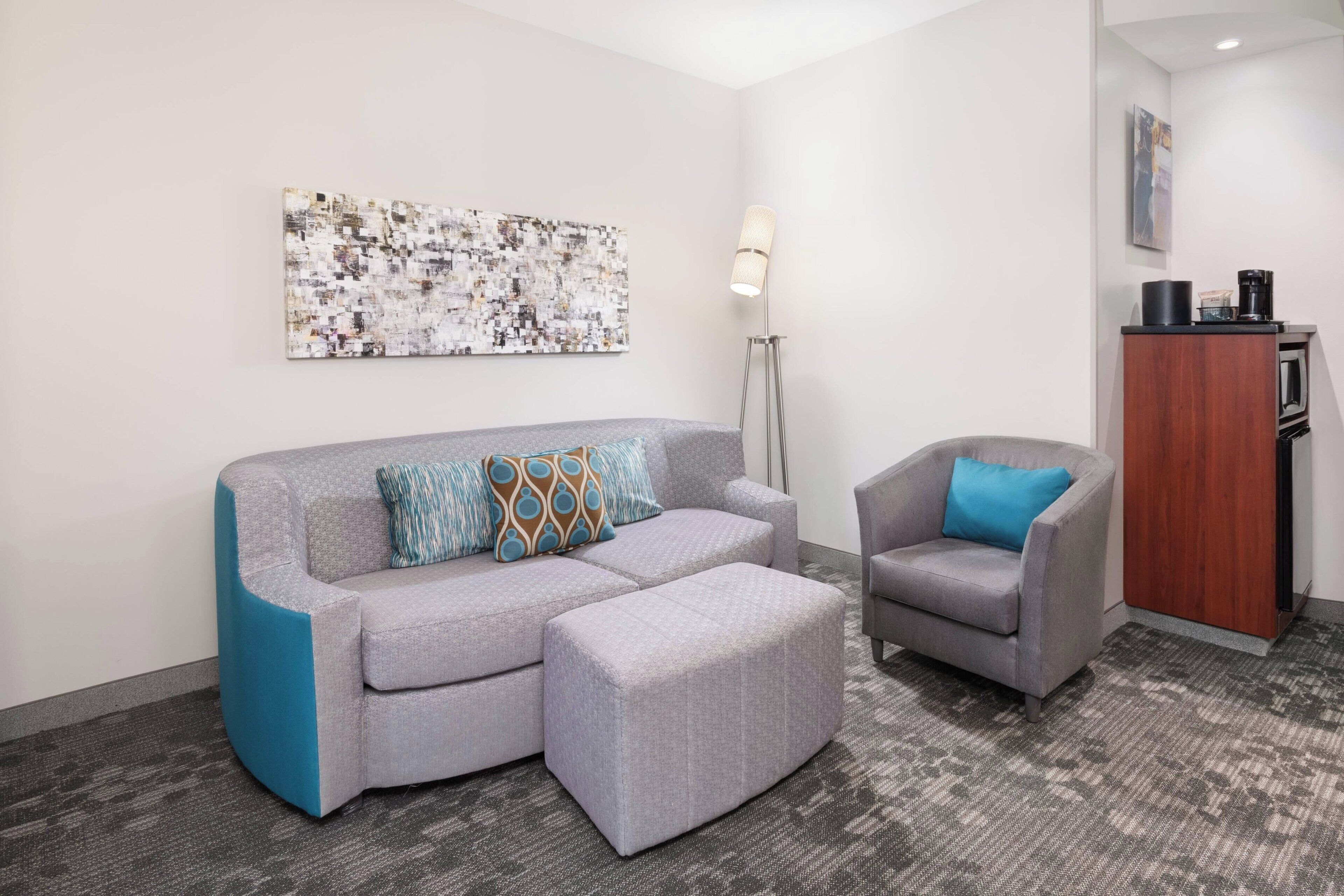 A modern living room with a grey sectional sofa, a matching ottoman, and a grey armchair with a blue pillow. A floor lamp and a wooden cabinet with a coffee machine are also visible.