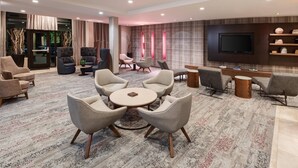 A modern lounge area with a round table surrounded by chairs, a flat-screen TV mounted on the wall, and a bookshelf with decorative items. - Courtyard by Marriott Pittsburgh Washington/Meadow Lands (Washington)