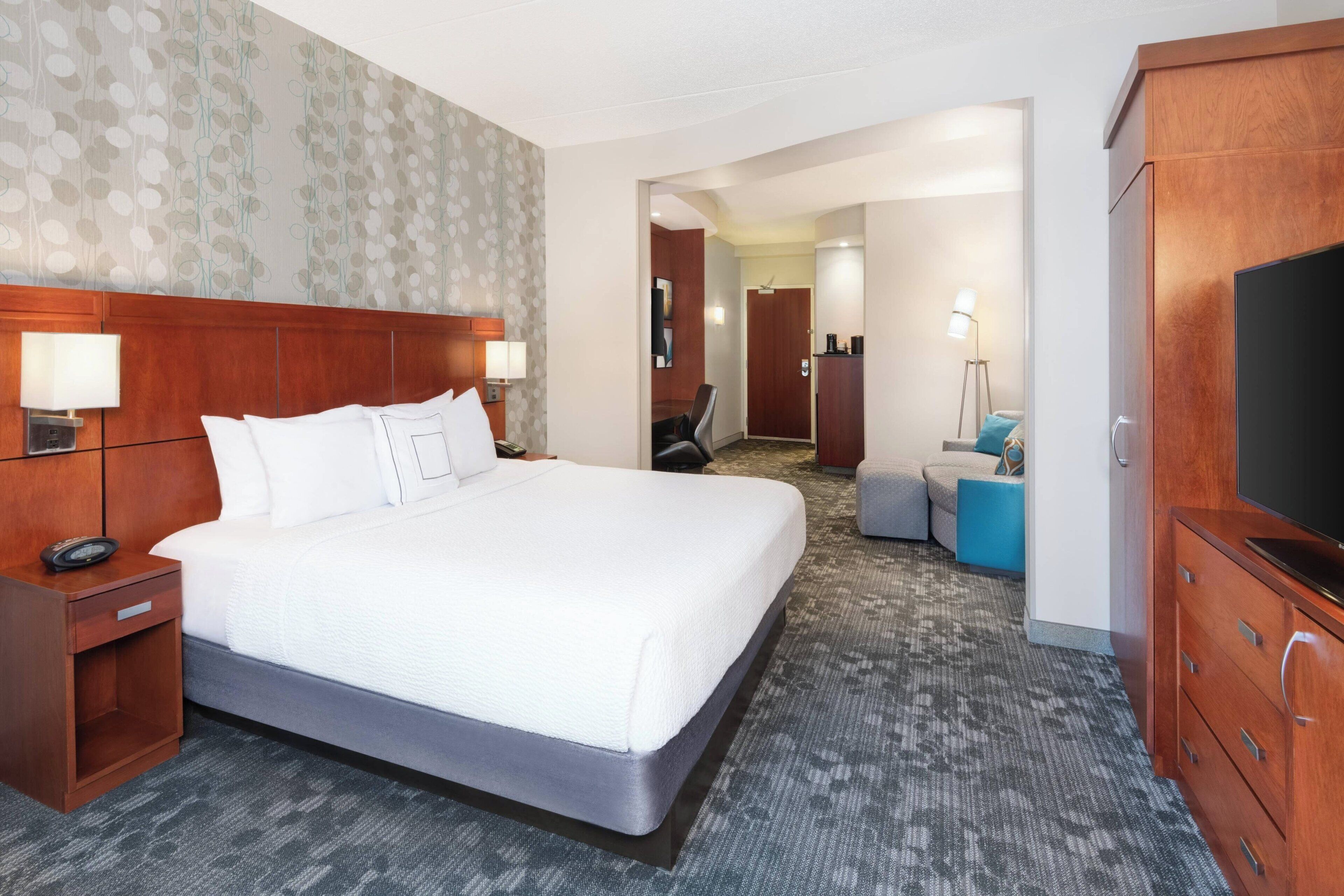 A hotel room with a large bed, a wooden headboard, a desk with a lamp, a television, and a seating area with a chair and ottoman.