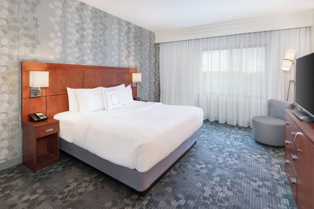 Une chambre d’hôtel avec un grand lit, une tête de lit en bois, une table de chevet avec un téléphone, un téléviseur posé sur une commode en bois et une fenêtre avec des rideaux.