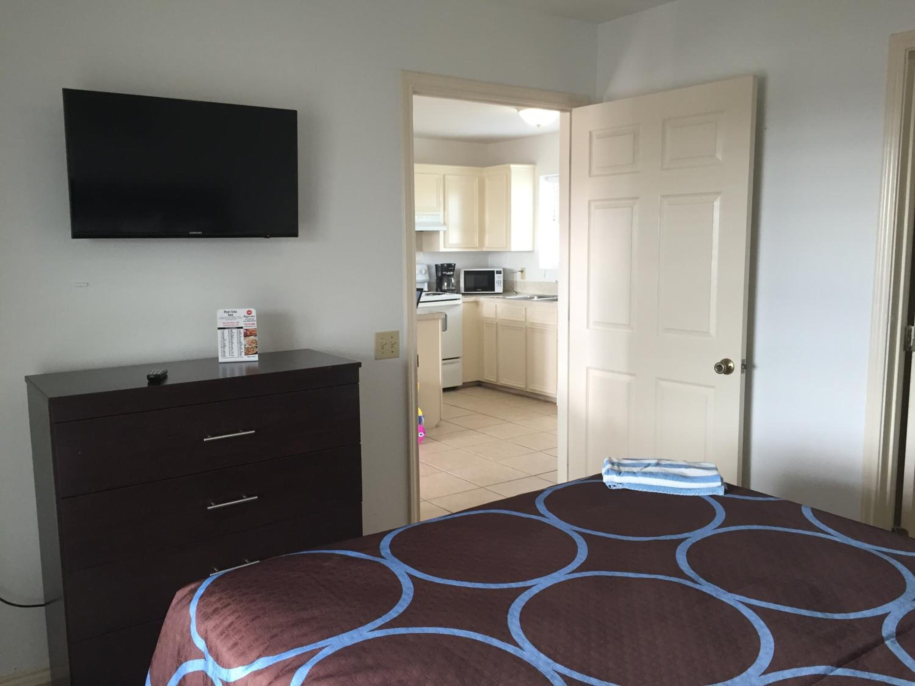A bedroom with a bed, a dresser, a television, and a view into a kitchen.
