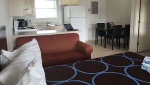 A compact hotel room with a bed, a sofa, a dining table with chairs, a kitchenette, and a small window.