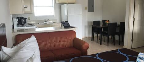 Una habitación de hotel compacta con cama, sofá, mesa de comedor con sillas, kitchenette y un ventanal pequeño.