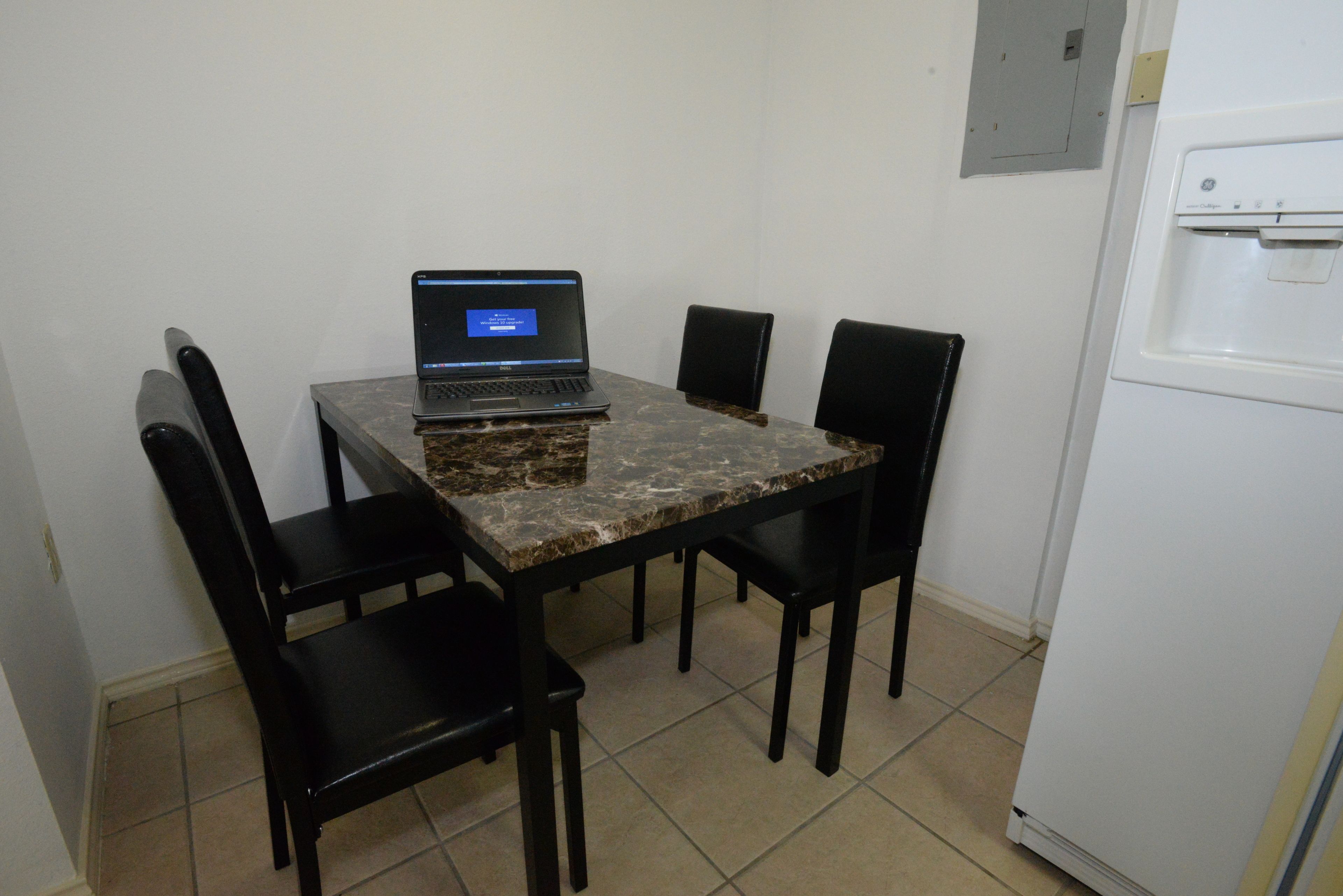 A small room with a tiled floor, a marble-top table, four black chairs, a laptop on the table, and a white refrigerator.