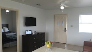 A hotel room with a bed, a desk with a laptop, a TV, a fruit bowl, and a ceiling fan.