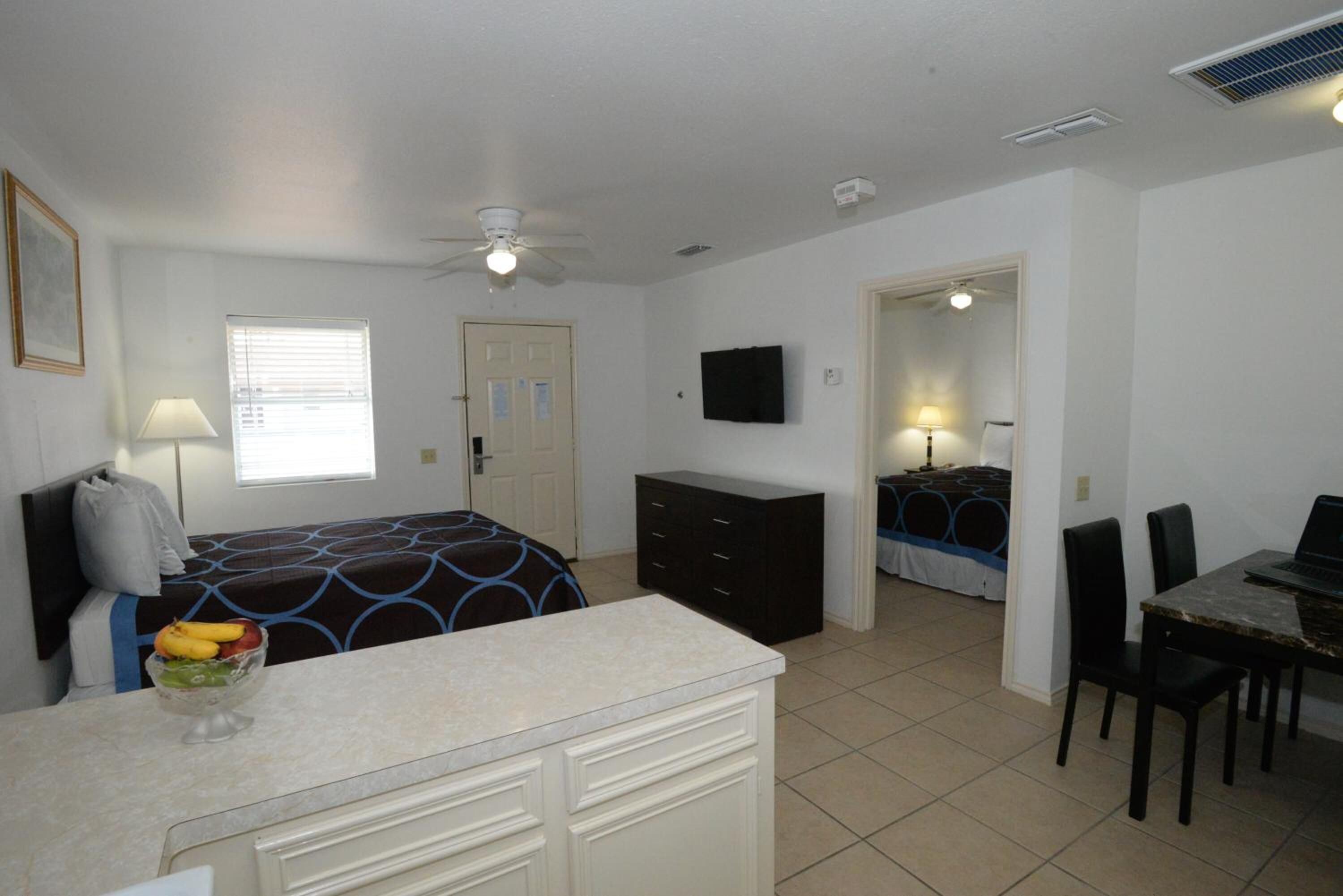 A bedroom with a bed, a dresser, a desk, and a chair. There is a window with blinds, a ceiling fan, and a television on the wall.