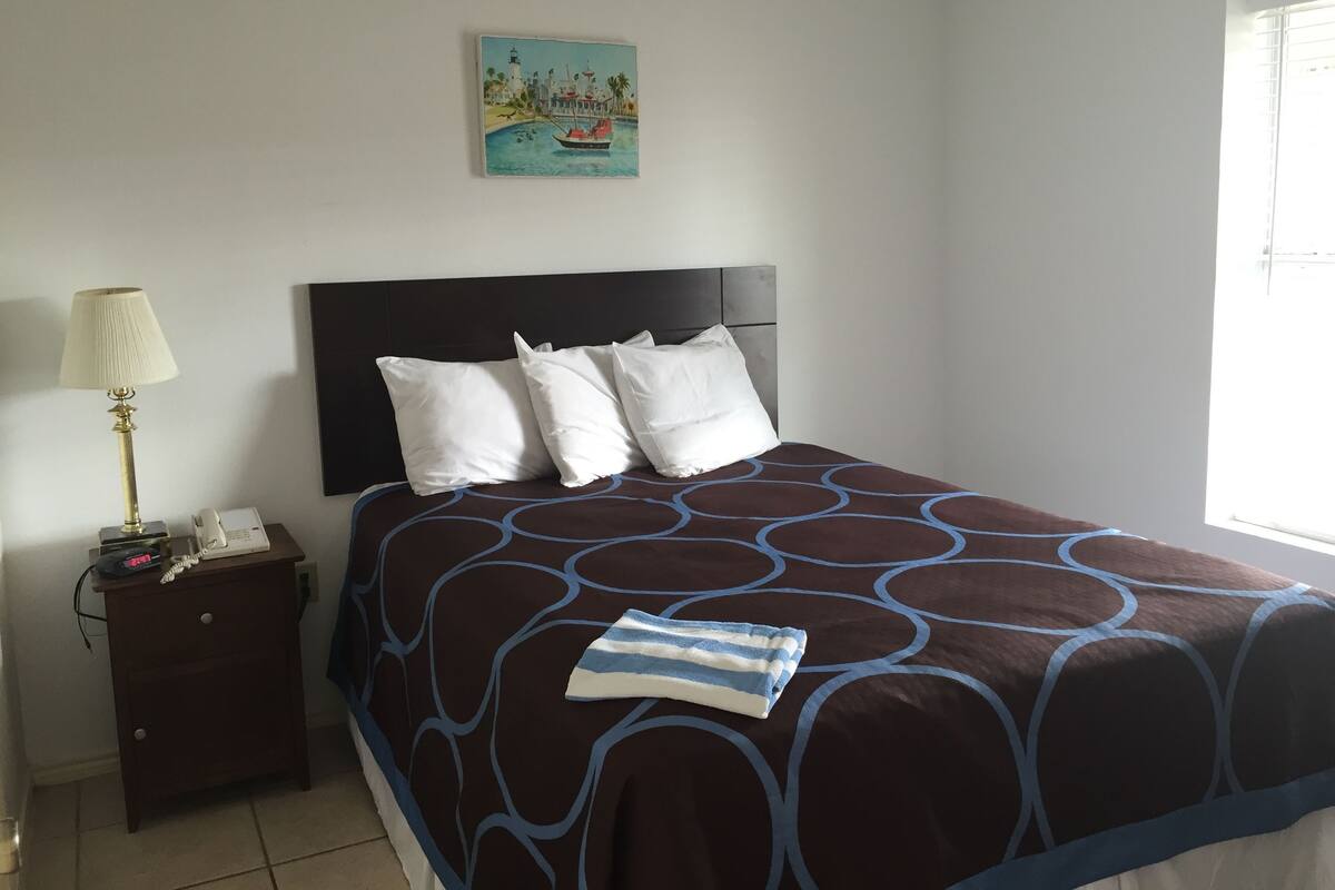 A neatly made bed with a patterned bedspread, a bedside lamp, a nightstand with a phone, and a painting of a coastal scene above the bed.