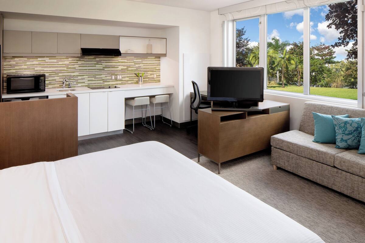 A modern hotel room with a large bed, a kitchenette, a TV, and a sofa with cushions.