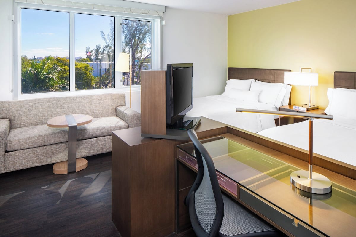 A hotel room with a bed, a desk, a television, a sofa, and a window with a view of trees and buildings.