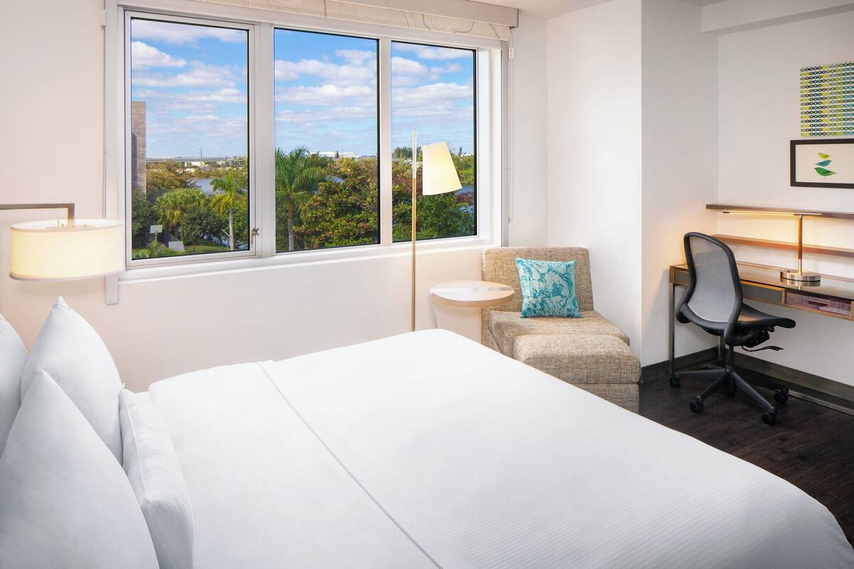 A hotel room with a large bed, a desk with a chair, a small round table, and a view of greenery through the window.