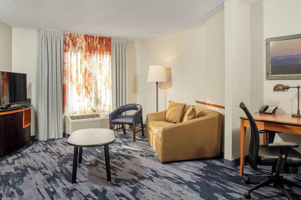 A hotel room with a sofa, a desk, a chair, a TV, and a lamp.