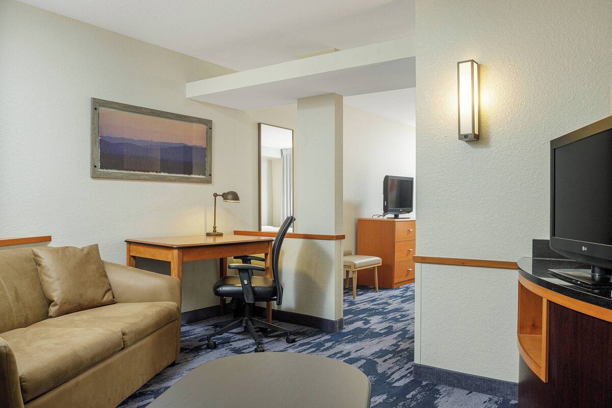 A hotel room with a sofa, desk, chair, television, and a painting on the wall.