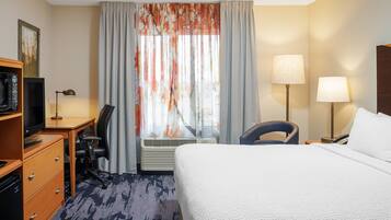 A hotel room with a bed, desk, chair, television, air conditioning unit, and a lamp.