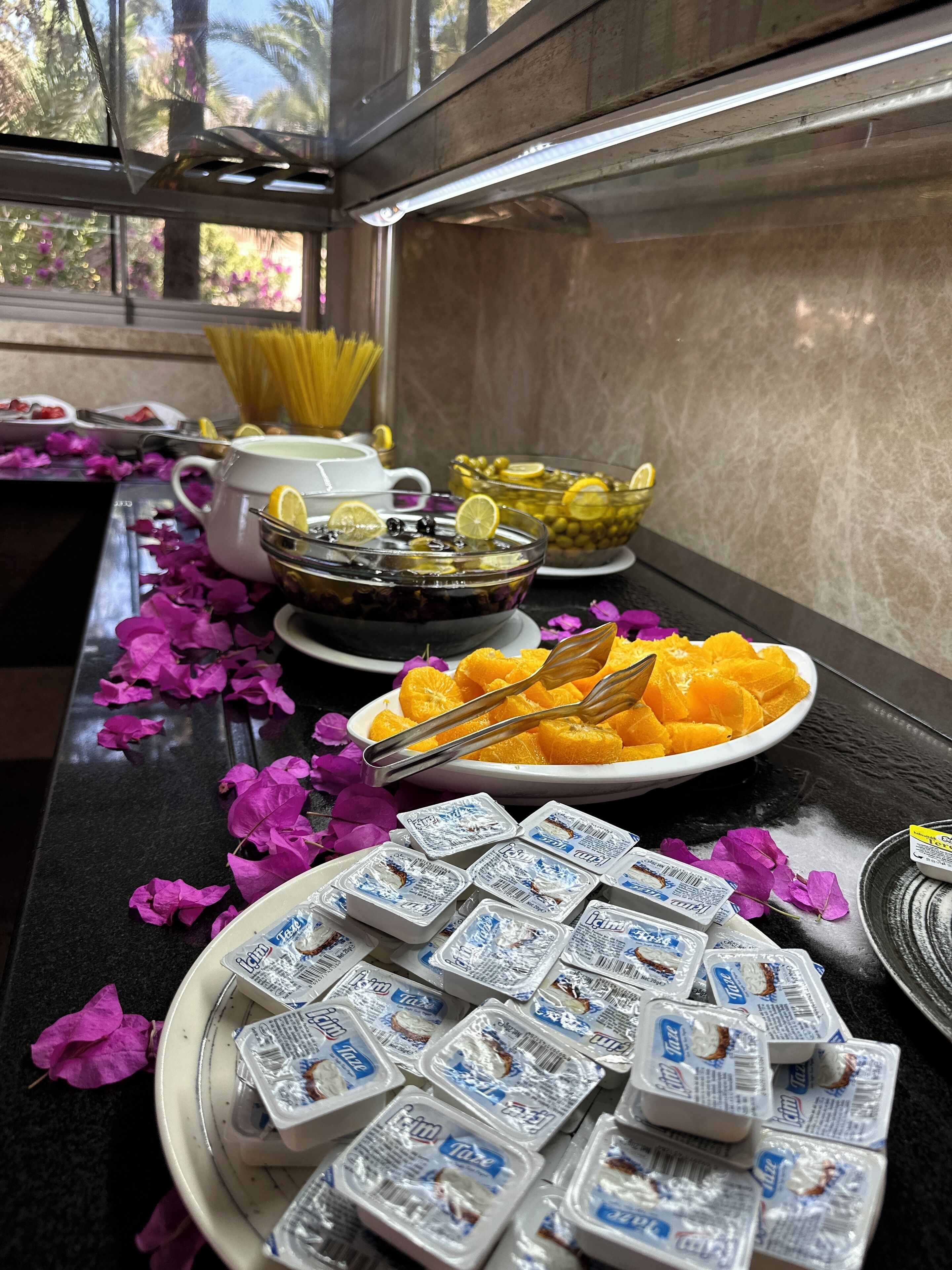 A buffet table with various food items including pasta, lemon slices, olives, and orange segments, along with packaged yogurt.