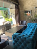 Uma sala de estar moderna com um sofá azul estofado, uma mesa de centro de madeira e uma poltrona preta. Um quadro está na parede, e há vista para o verde lá fora.