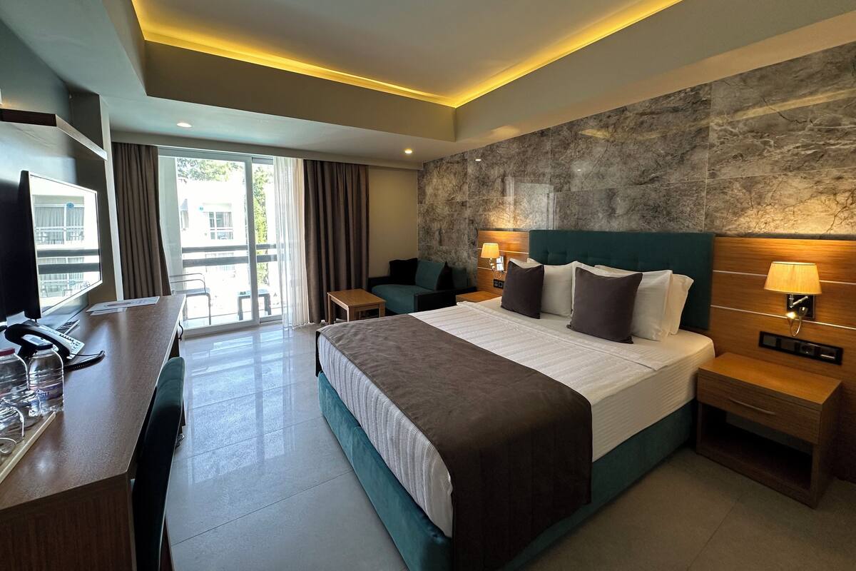 Quarto de hotel moderno com uma cama grande, uma escrivaninha com computador e telefone, um espaço de estar e vista pela porta de vidro.
