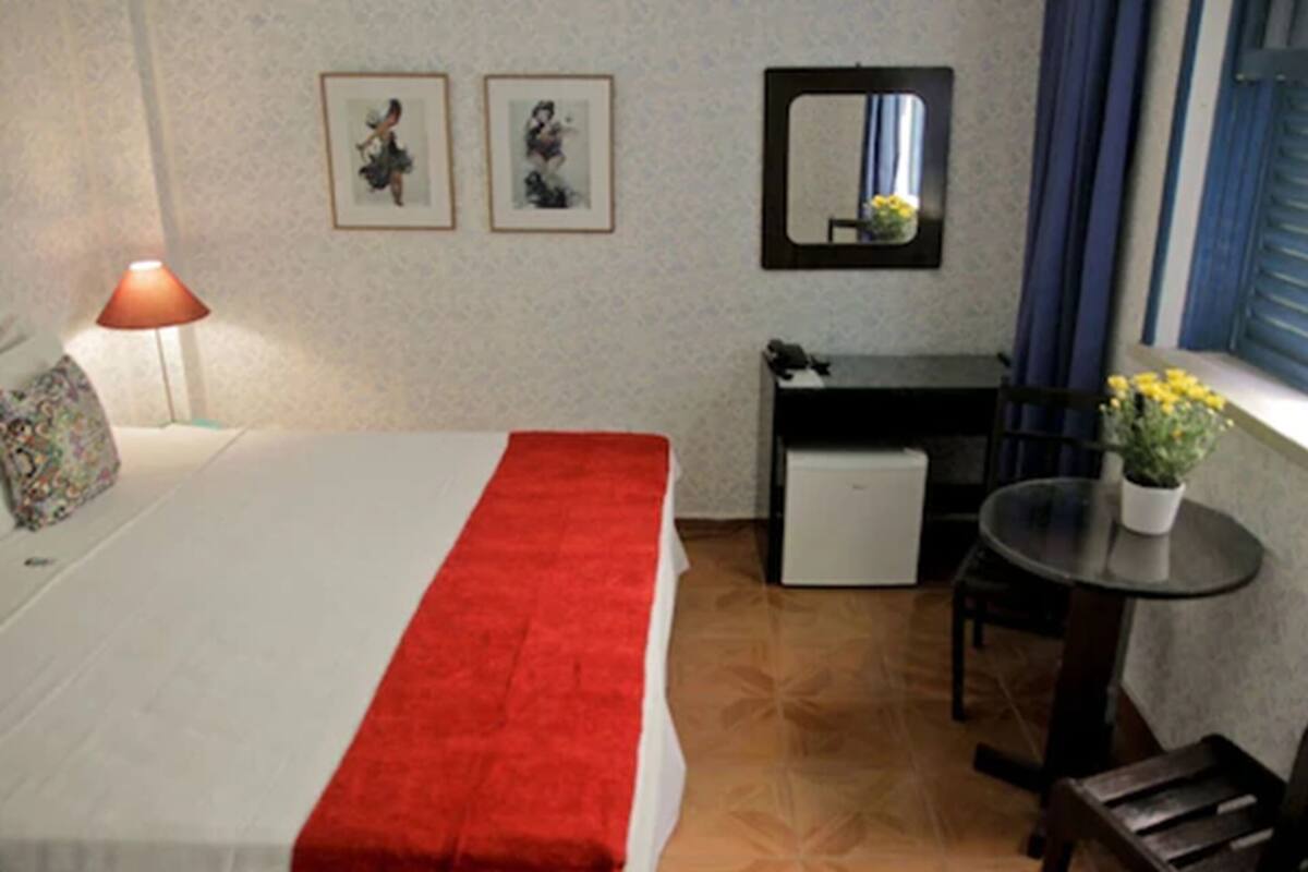 Habitació d'hotel amb un llit, escriptori, cadira, taula petita amb una planta, làmpada i finestra amb cortines.
