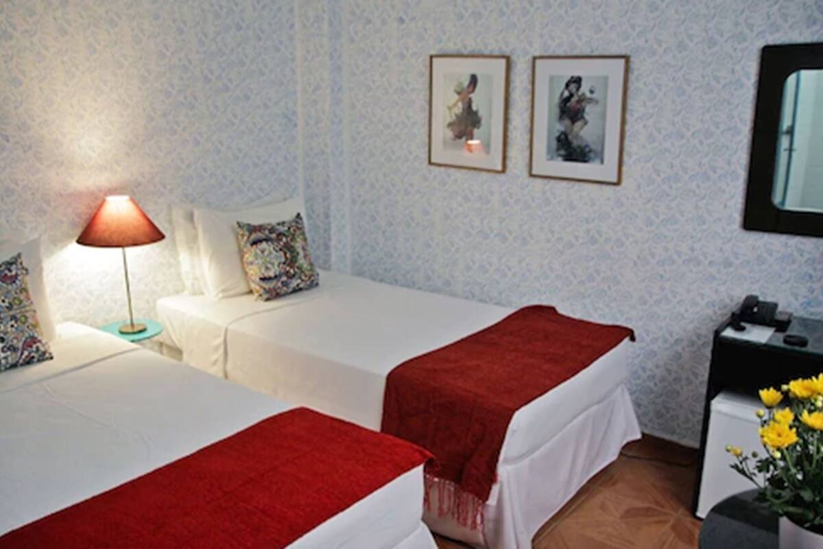 Habitació d'hotel amb dos llits, una tauleta de nit, una làmpada i dos quadres al mur.