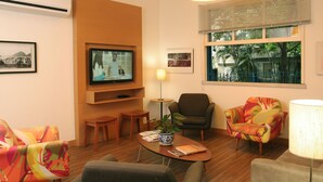 Una sala de estar moderna con una televisión de pantalla plana, asientos cómodos, una mesa de centro y un ventanal con persianas.