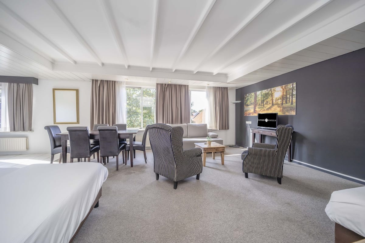 Une chambre d’hôtel comprenant un lit, deux fauteuils à rayures, un canapé, une table à manger avec des chaises, un téléviseur et de grandes fenêtres avec des rideaux.
