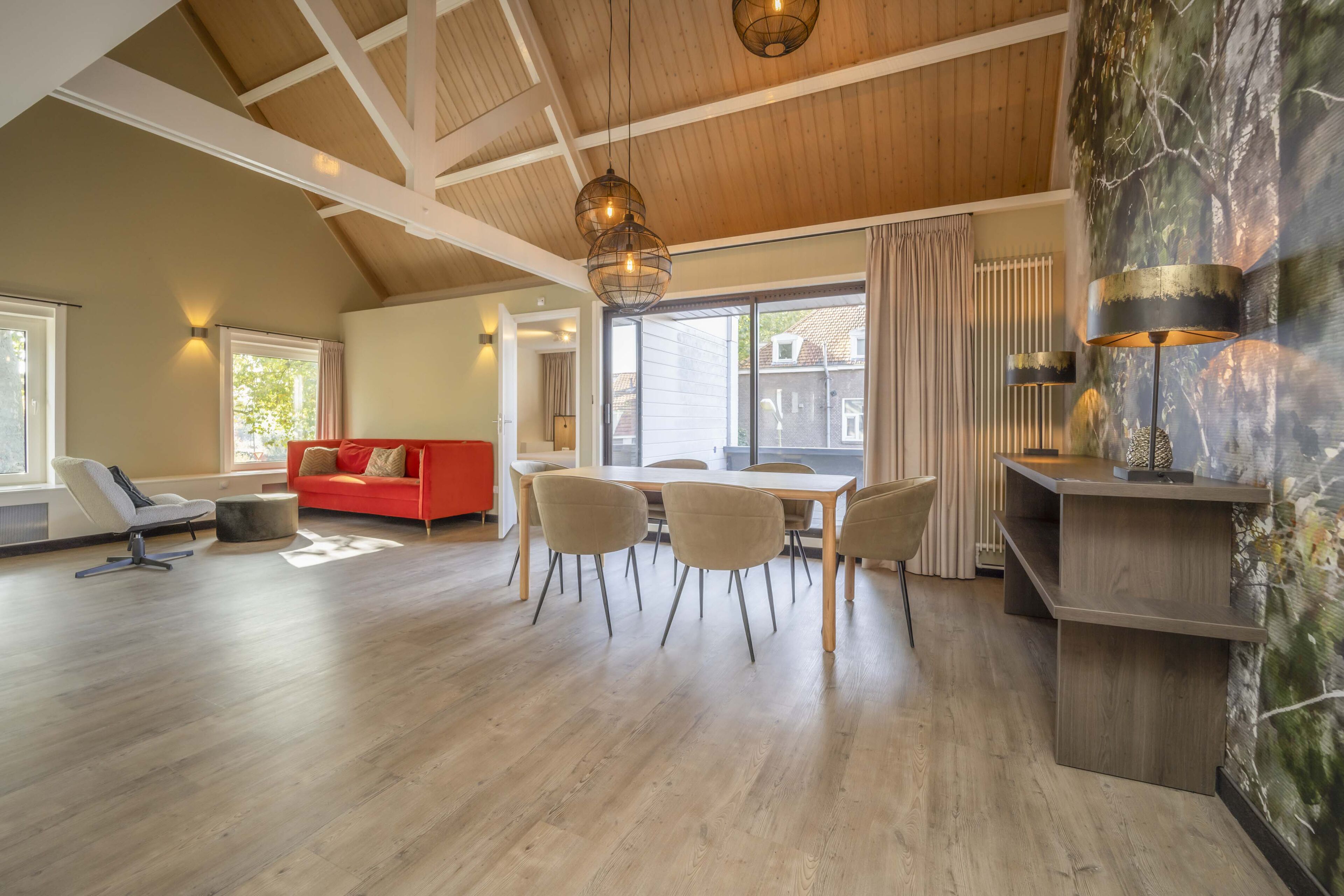 A spacious living area with a high ceiling, a dining table with chairs, a red sofa, and a large window with curtains.