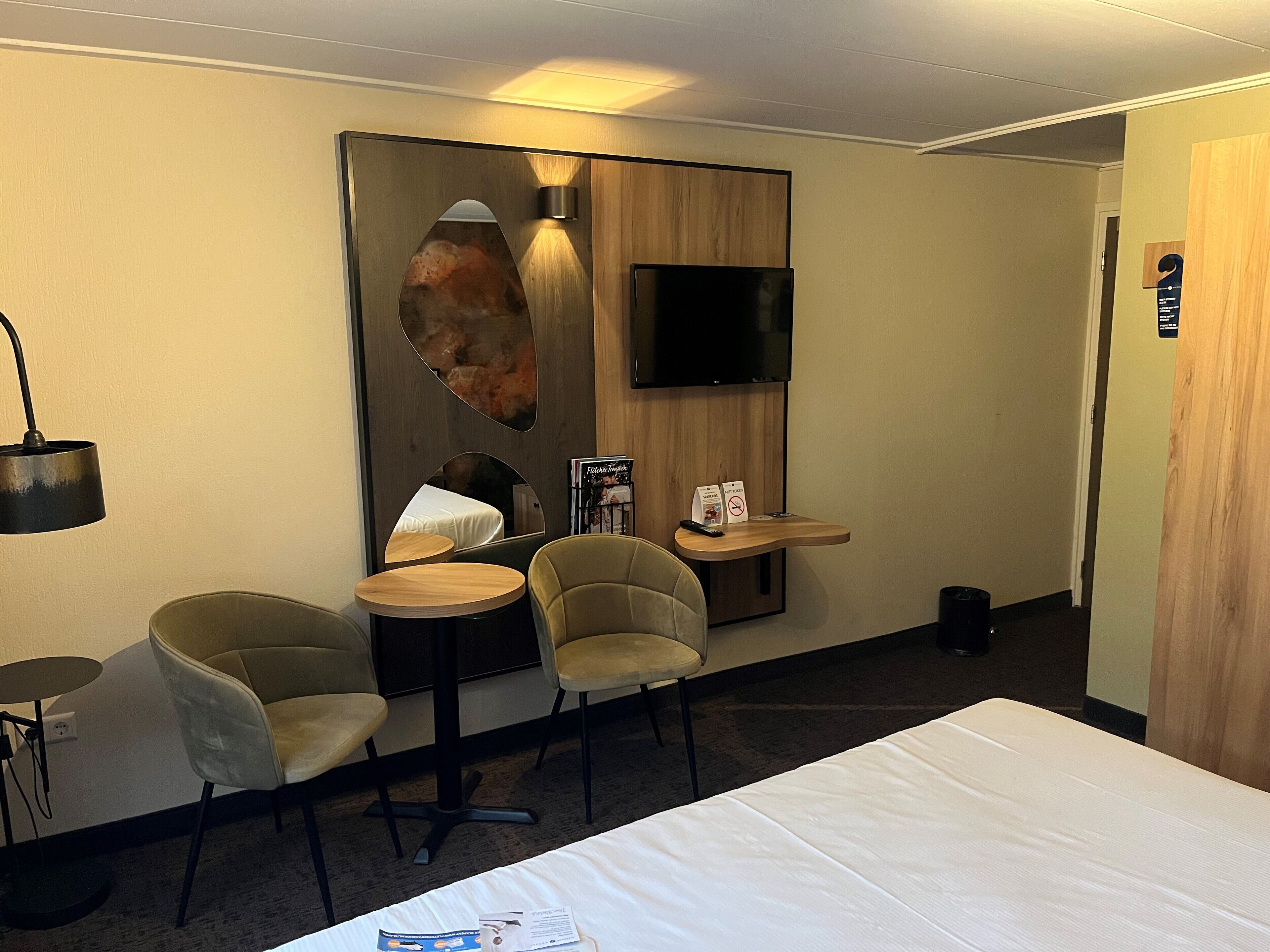 A hotel room with a bed, two chairs, a small table, a TV, and a mirror.