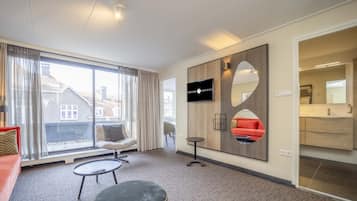 A modern hotel room with a large window, a flat-screen TV, a sofa, a coffee table, and a small round table.