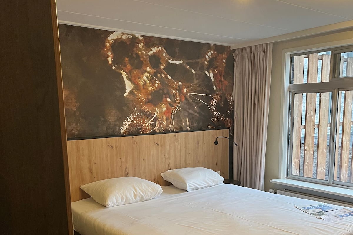 Une chambre d’hôtel moderne avec un grand lit, une tête de lit en bois, un grand tableau abstrait au mur et une fenêtre avec des rideaux.