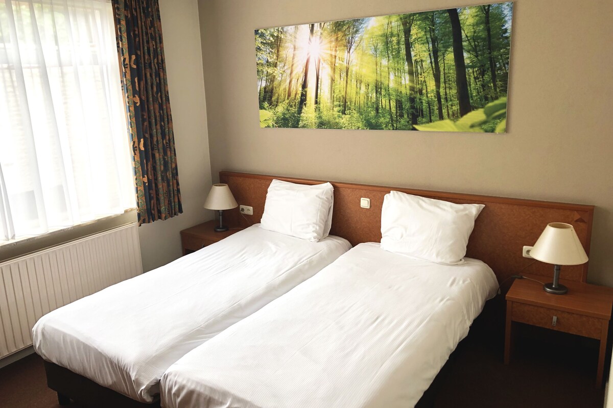 A double bed with white linens, two bedside tables with lamps, a window with sheer curtains, and a forest-themed painting on the wall.