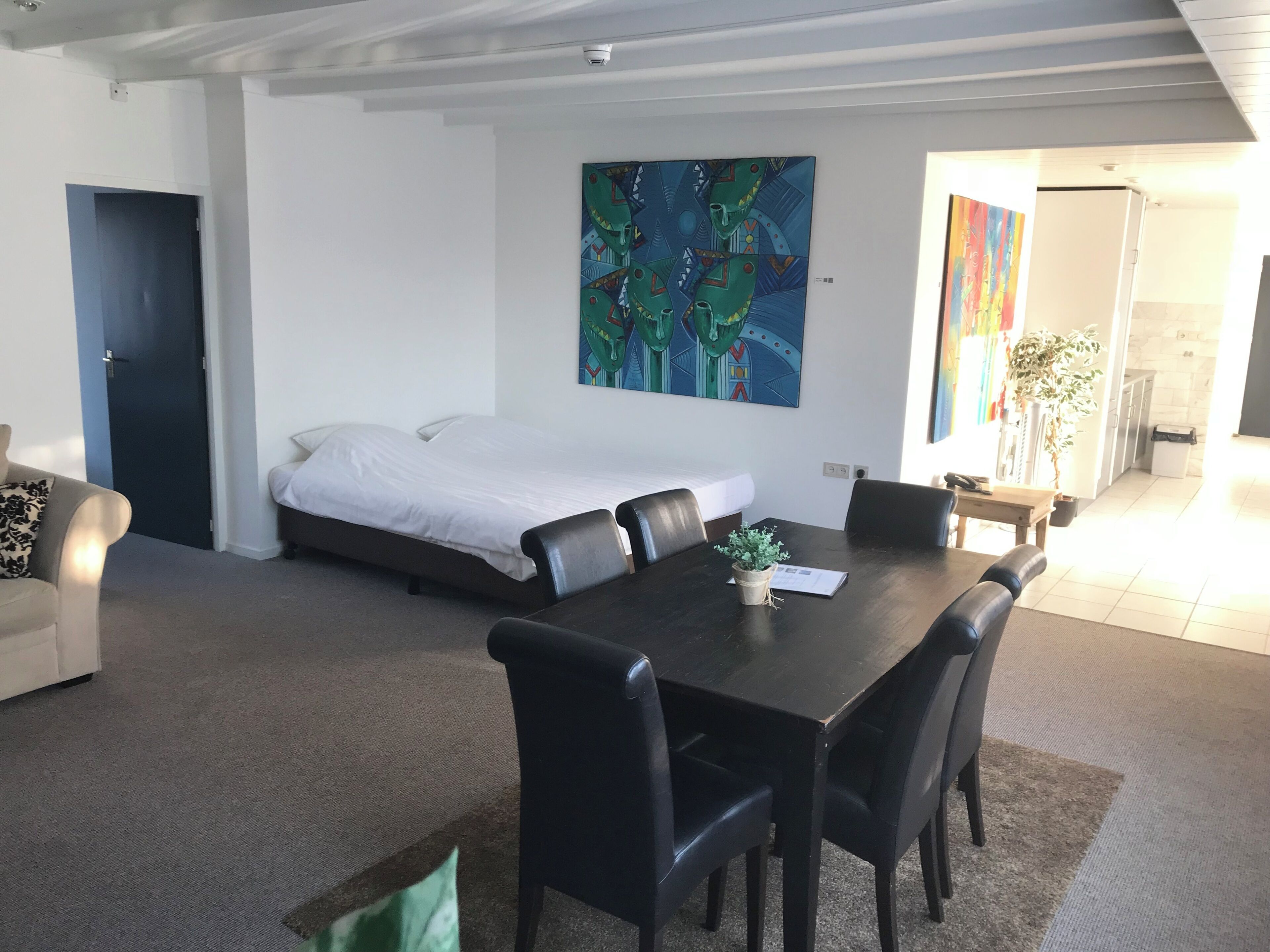 A modern hotel room with a bed, a dining table with chairs, a small potted plant, and a painting on the wall.