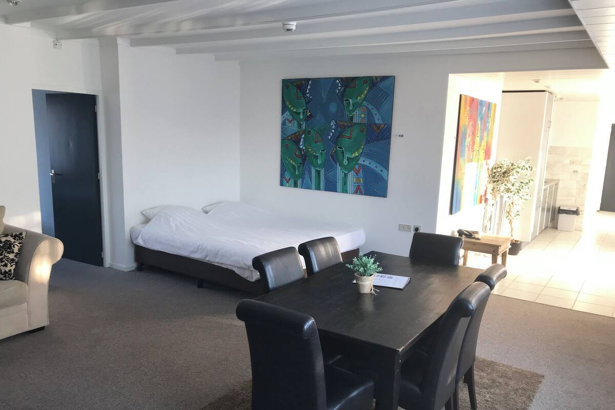 Une chambre d’hôtel moderne, avec un lit, une table à manger et des chaises, une petite plante en pot et un tableau accroché au mur.