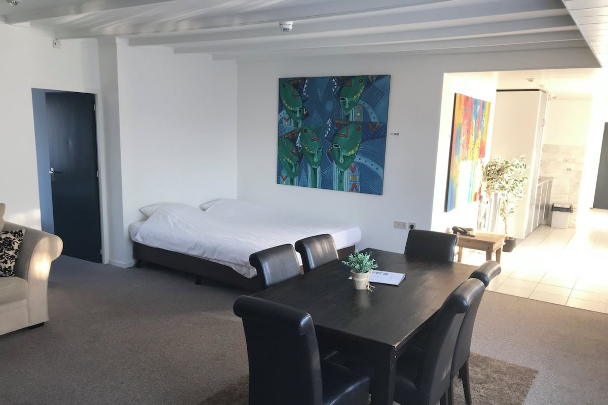 A modern hotel room with a bed, a dining table with chairs, a small potted plant, and a painting on the wall.