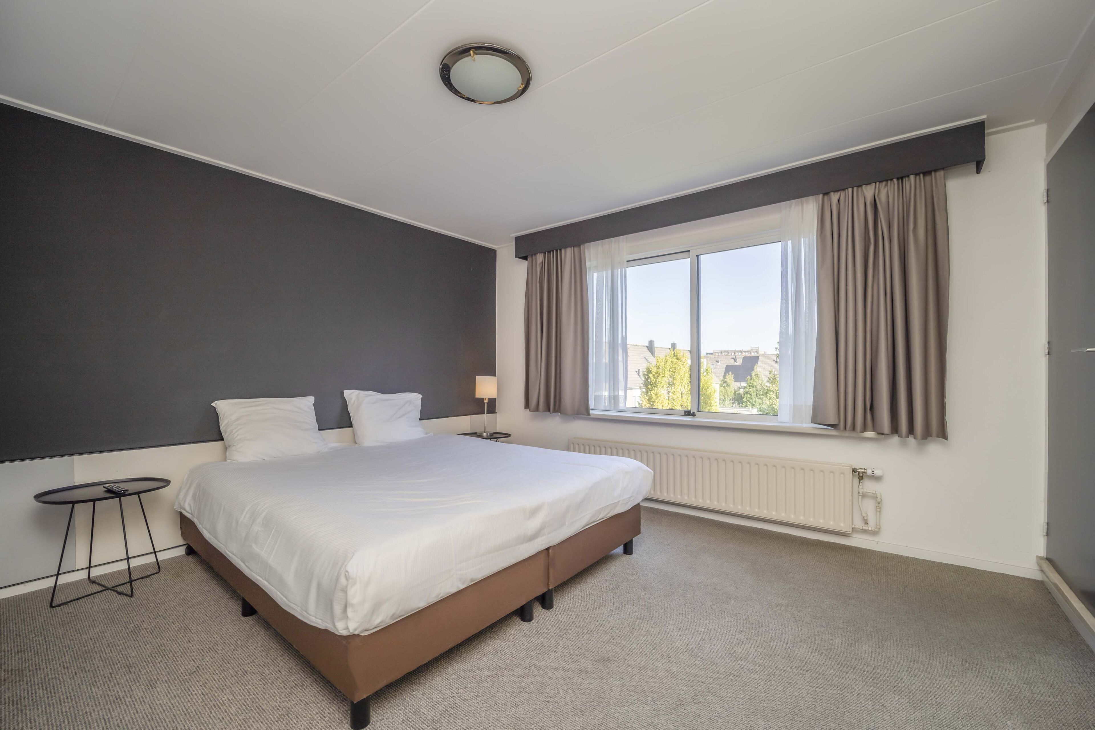 Une chambre d’hôtel avec un grand lit, une petite table, une fenêtre avec des rideaux et un radiateur.