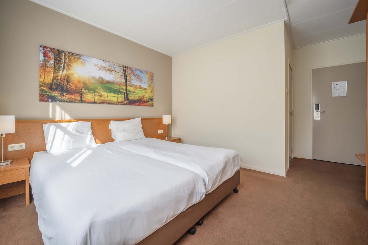 Une chambre d’hôtel avec un grand lit, deux tables de chevet avec des lampes, un tableau représentant une scène de forêt et une porte avec une serrure.