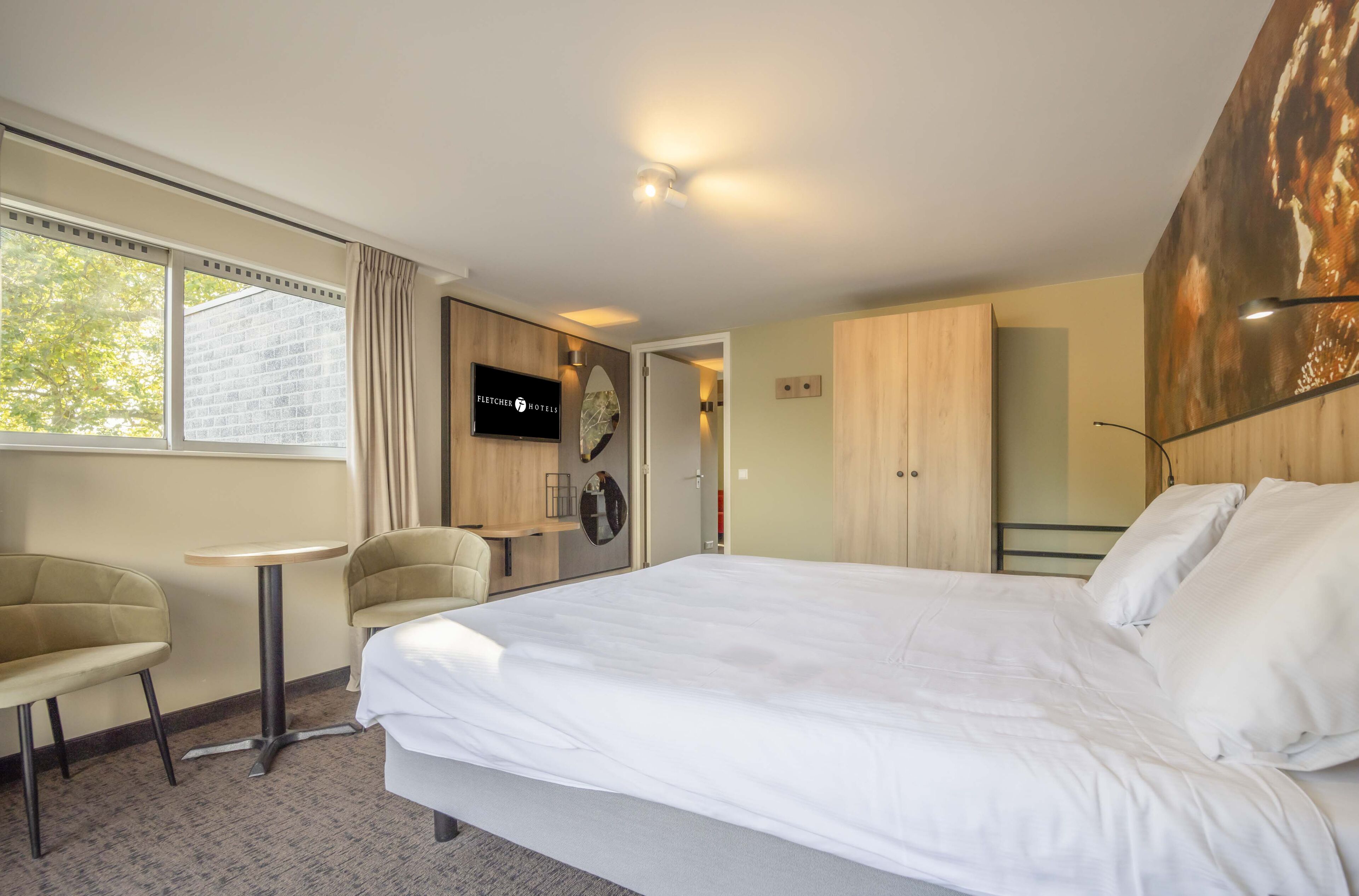 Quarto de hotel com cama, escrivaninha, duas cadeiras, televisão e uma janela grande.