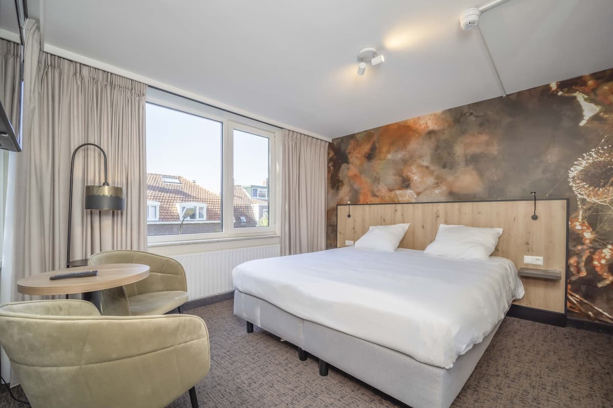Une chambre d’hôtel avec un grand lit, une table ronde, une chaise et une vue sur les bâtiments à travers la fenêtre.