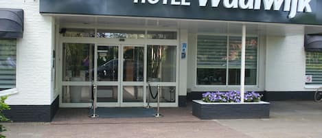 Entrada do HOTEL Waalwijk com porta de vidro, barreira de metal e canteiro de flores roxas.