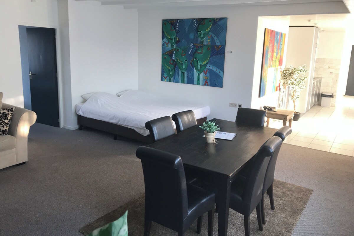 A modern hotel room with a bed, a dining table with chairs, a small potted plant, and a painting on the wall.