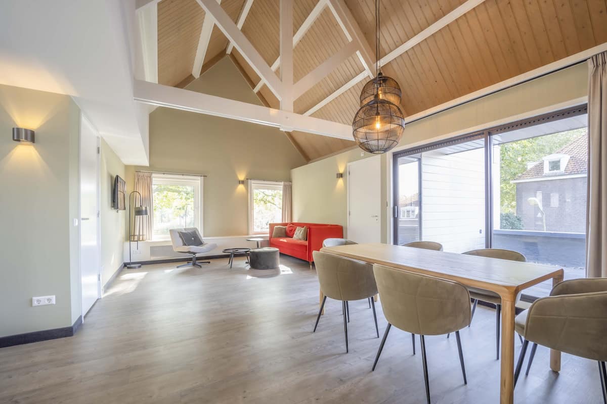 Un vaste espace de vie doté d’un plafond en bois, d’une table à manger avec des chaises, d’un canapé rouge et de grandes fenêtres laissant entrer la lumière naturelle.