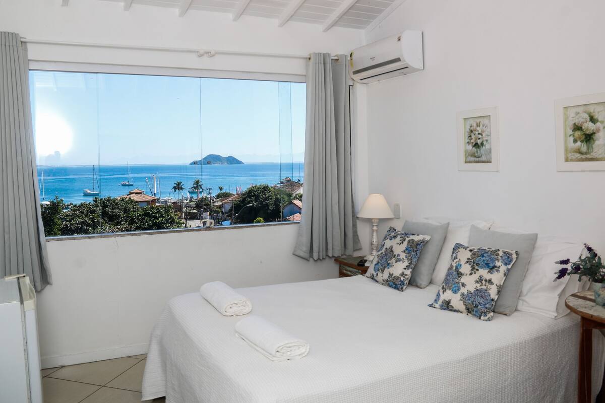 A bedroom with a large window overlooking the sea, a bed with white bedding, and a nightstand with a lamp.