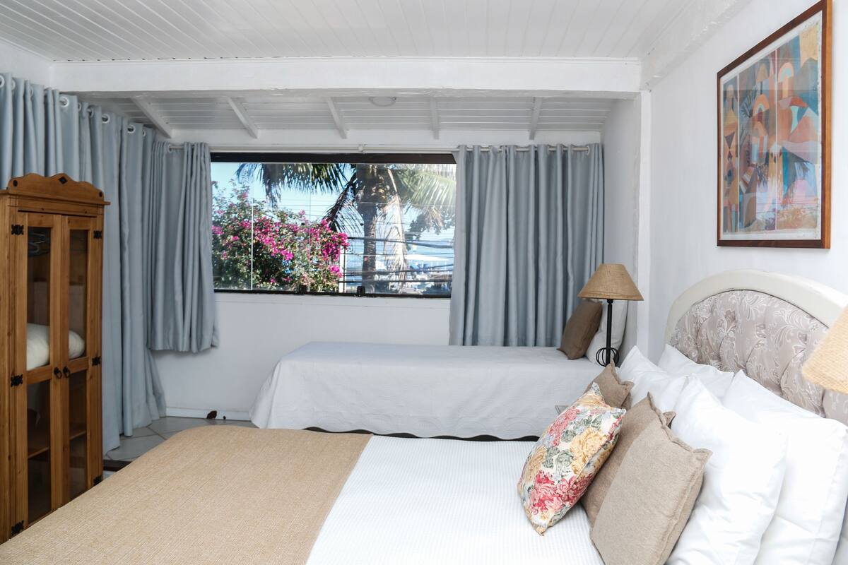 A bedroom with two beds, a wooden wardrobe, a window with a view of palm trees and flowers, and a painting on the wall.