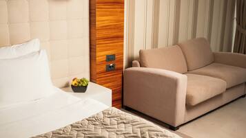 A hotel room with a bed, a beige sofa, a wooden accent wall, a nightstand with a fruit bowl, and a wall-mounted lamp.