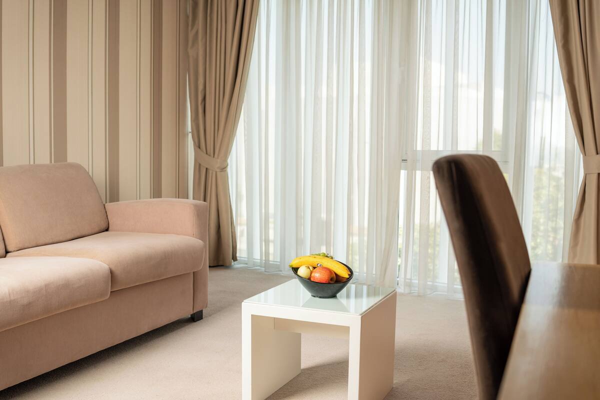 Una sala de estar moderna con un sofá color crema, una mesa de centro de vidrio con un frutero, y vistas a través de cortinas traslúcidas.