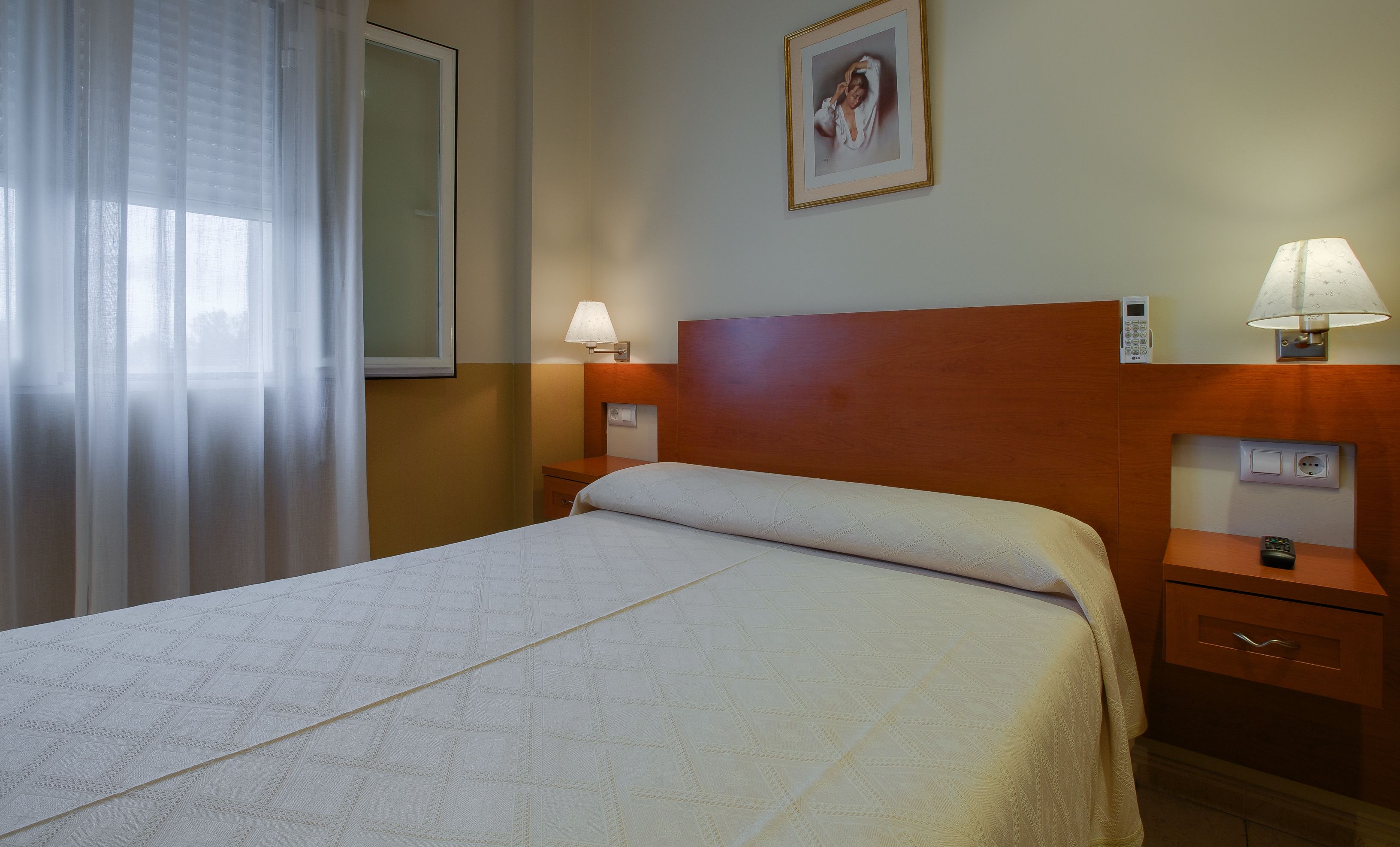 Habitación de hotel con cama, dos mesitas de noche, lámpara, un cuadro en la pared y ventana con cortinas.
