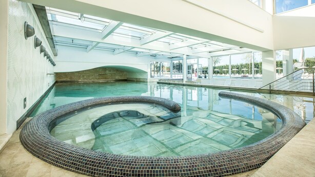 Une grande piscine intérieure avec un jacuzzi, le tout entouré d’un espace clos par des parois de verre, offrant une vue sur l’extérieur.