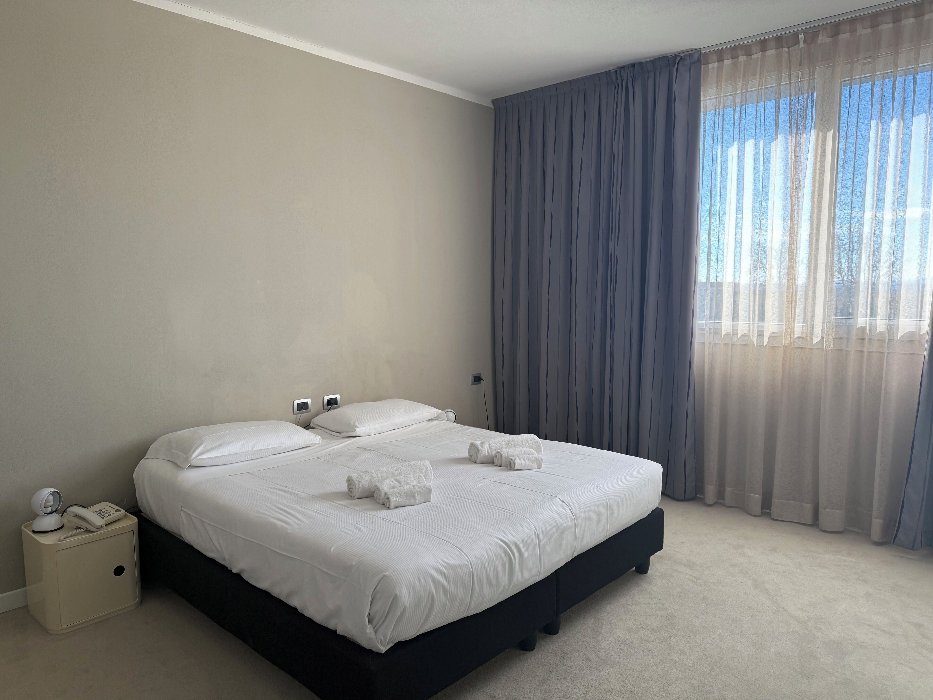 Una cama bien hecha con sábanas blancas y dos toallas enrolladas, una mesita de noche con un teléfono y una lámpara, y una ventana con cortinas de encaje.
