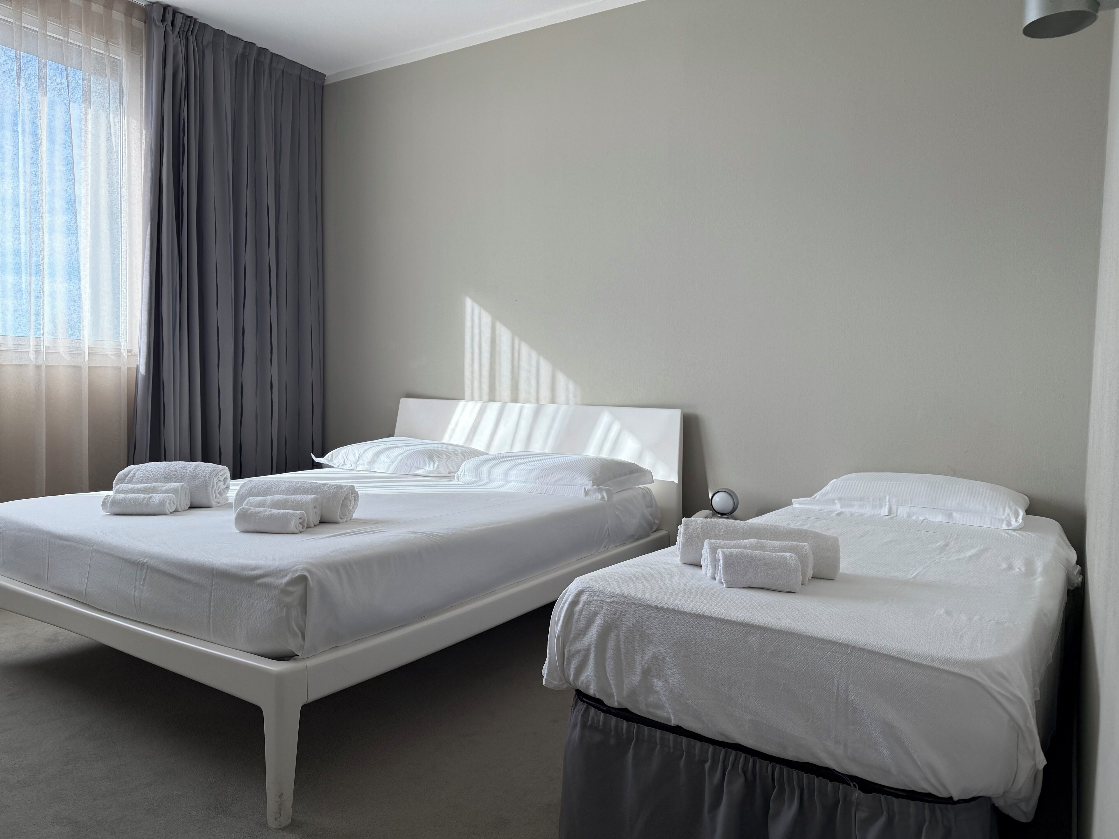 Una habitación de hotel con dos camas, un ventanal con cortinas translúcidas y una mesita de noche con lámpara.