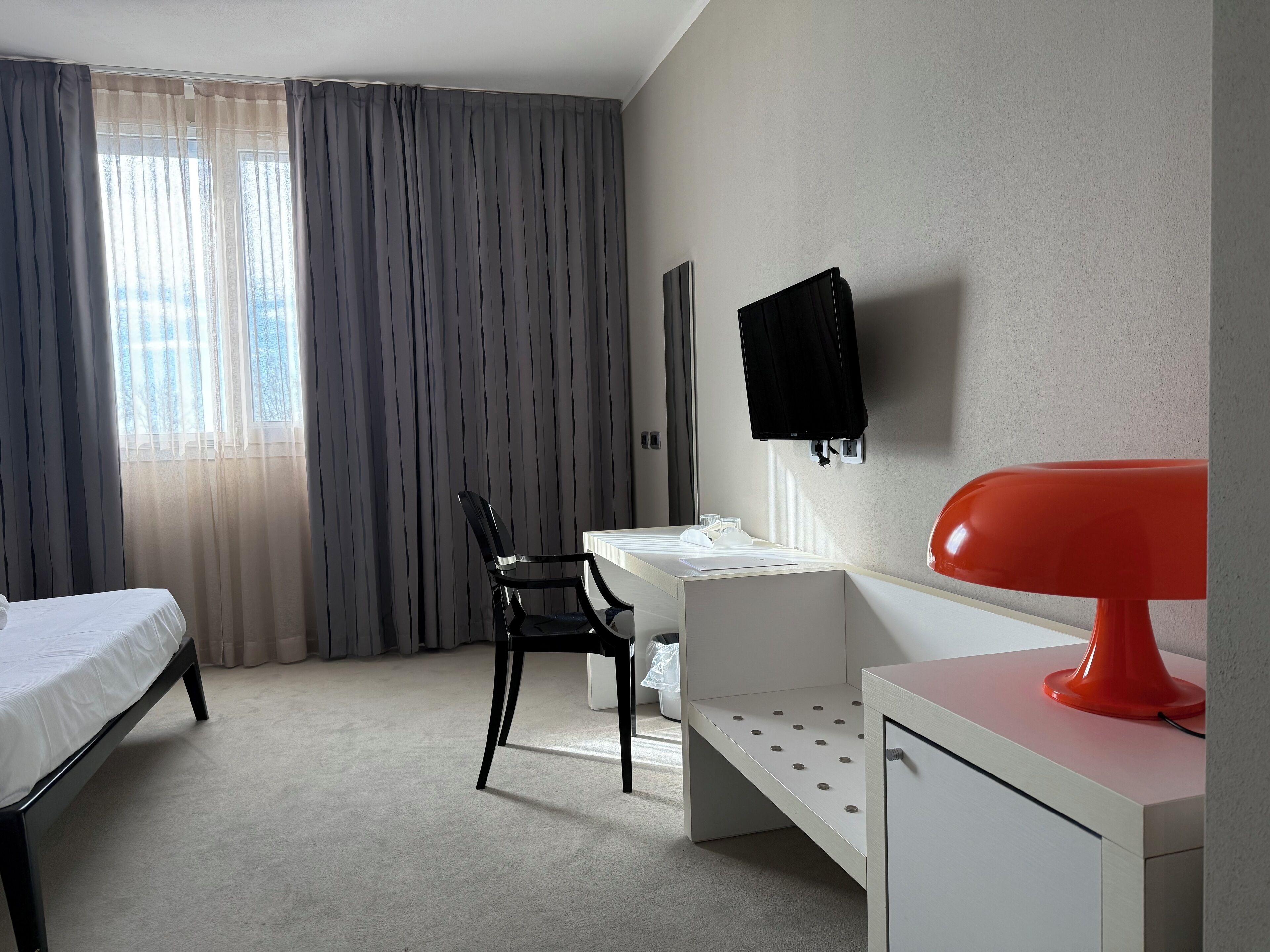 A hotel room with a bed, a desk, a chair, a TV mounted on the wall, and a bright red lamp.